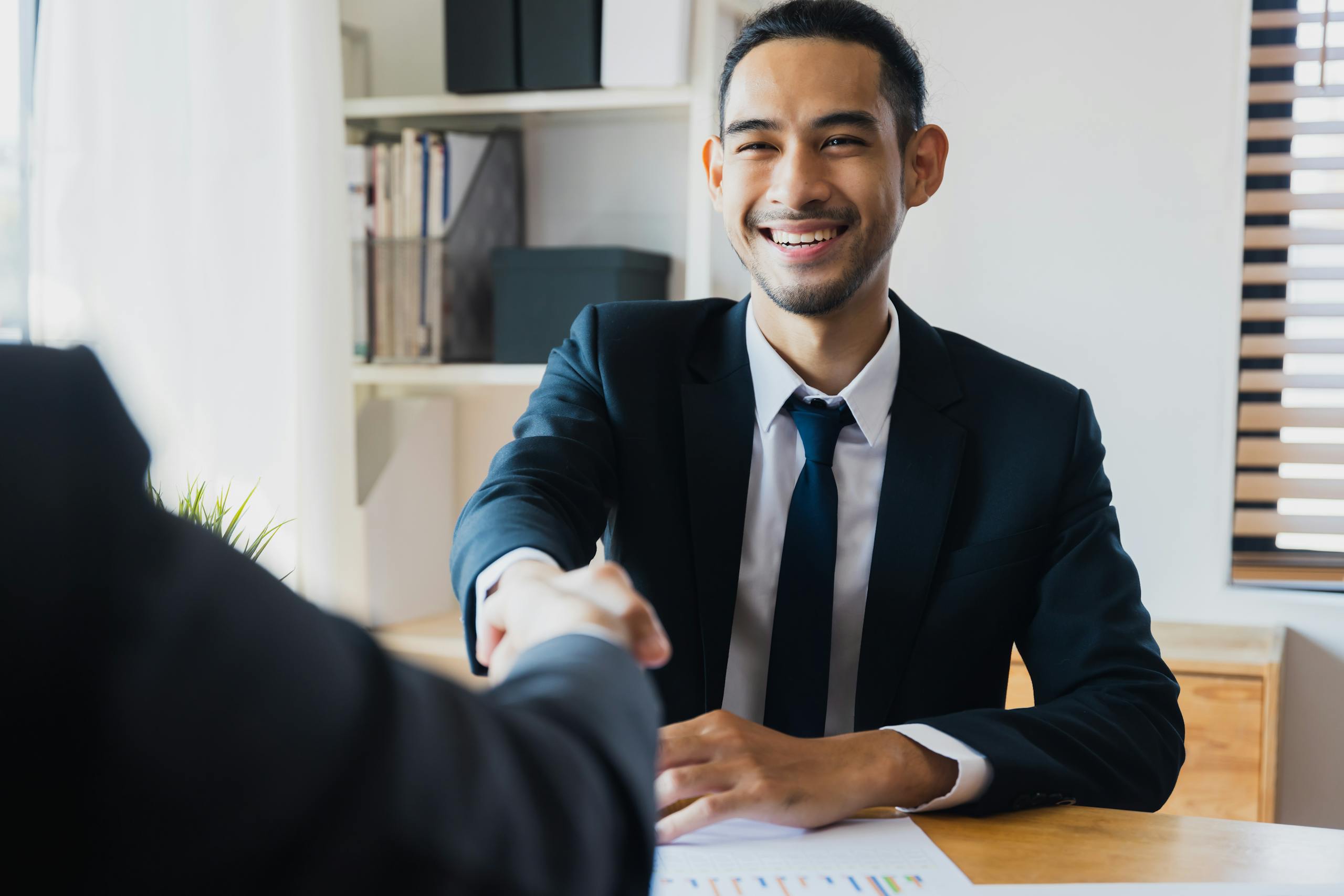 Confident businessman in suit shaking hands at office desk, symbolizing successful partnership after hiring a bookkeeper.